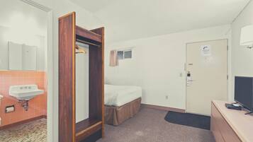 A hotel room with a wooden wardrobe, a single bed, a small window, a door with a sign, a sink, and a television on a dresser.