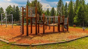 A playground with wooden climbing structures and a slide, surrounded by a wooded area. - Timbers at Island Park Village Resort (Island Park)