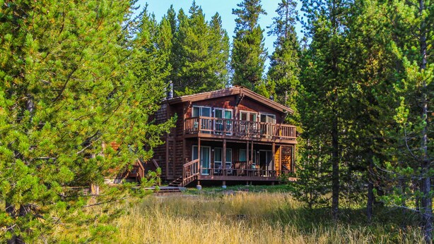 A two-story log cabin with a balcony, surrounded by dense forest. - Timbers at Island Park Village Resort (Island Park)