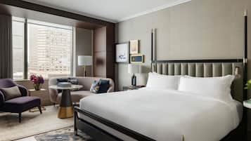 A hotel room with a large bed, a sofa, a small table, and a desk. There are two lamps, a vase with flowers, and a view of a building through the window.