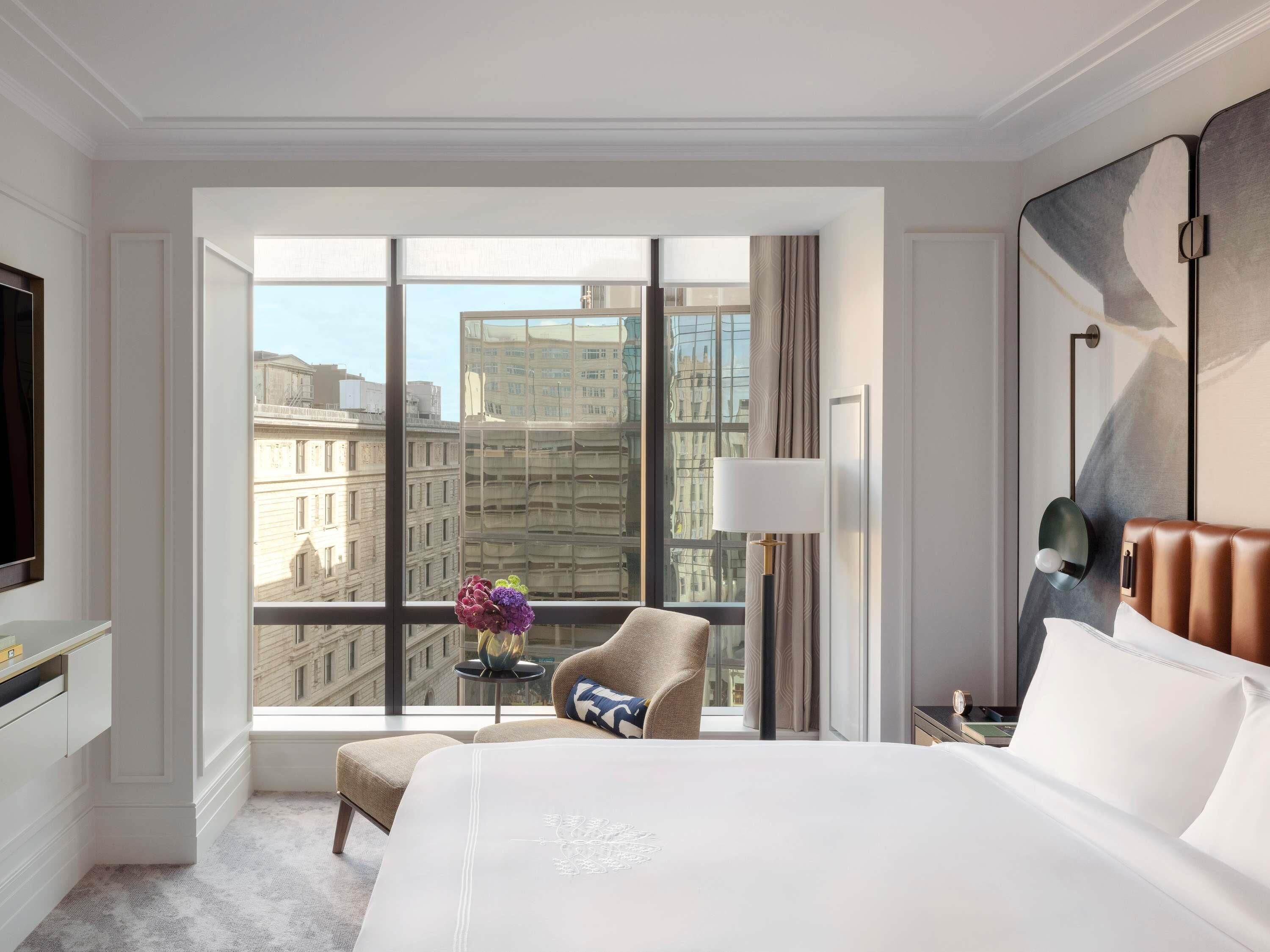 Une chambre d’hôtel avec une grande fenêtre, un lit, une chaise, une table de chevet et une lampe.