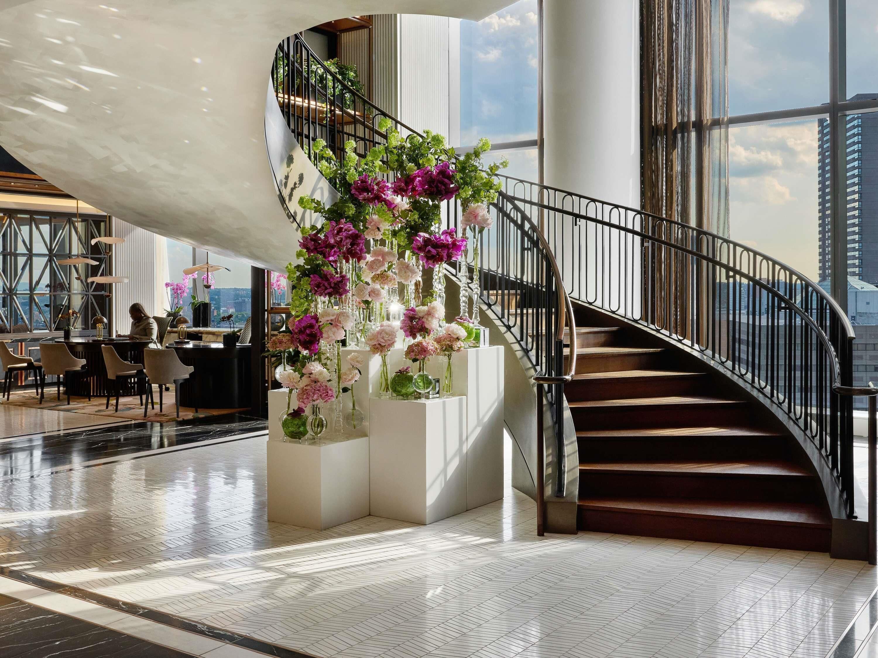 A modern interior with a curved staircase, a seating area with tables, and a view of the cityscape through large windows.