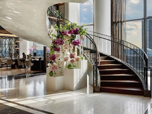 A modern interior with a curved staircase, a seating area with tables, and a view of the cityscape through large windows. - Raffles Boston (Boston)