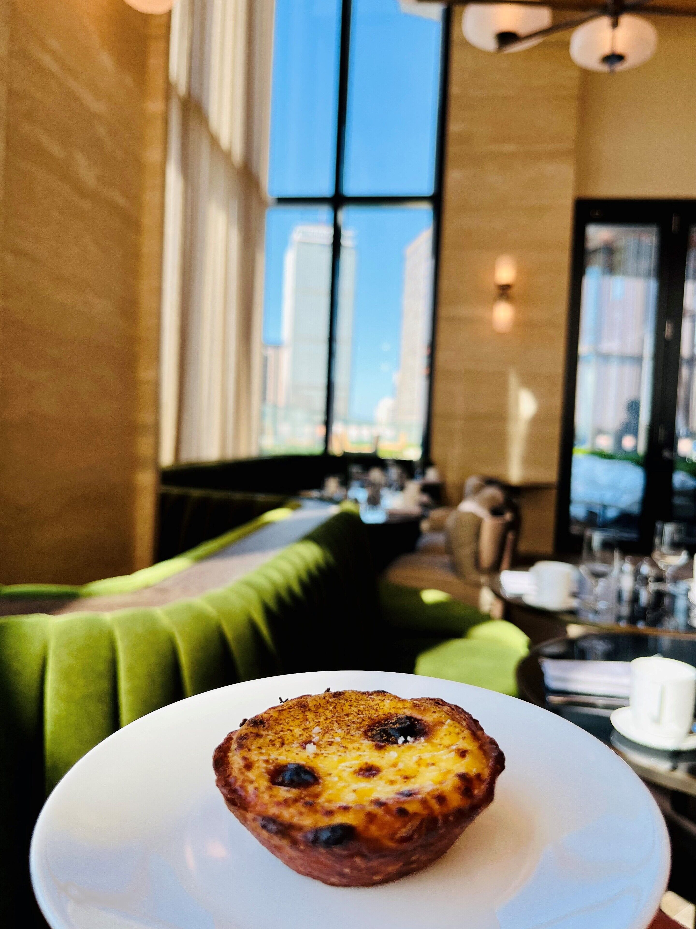 A muffin on a white plate in a restaurant with a green cushioned bench and large windows.