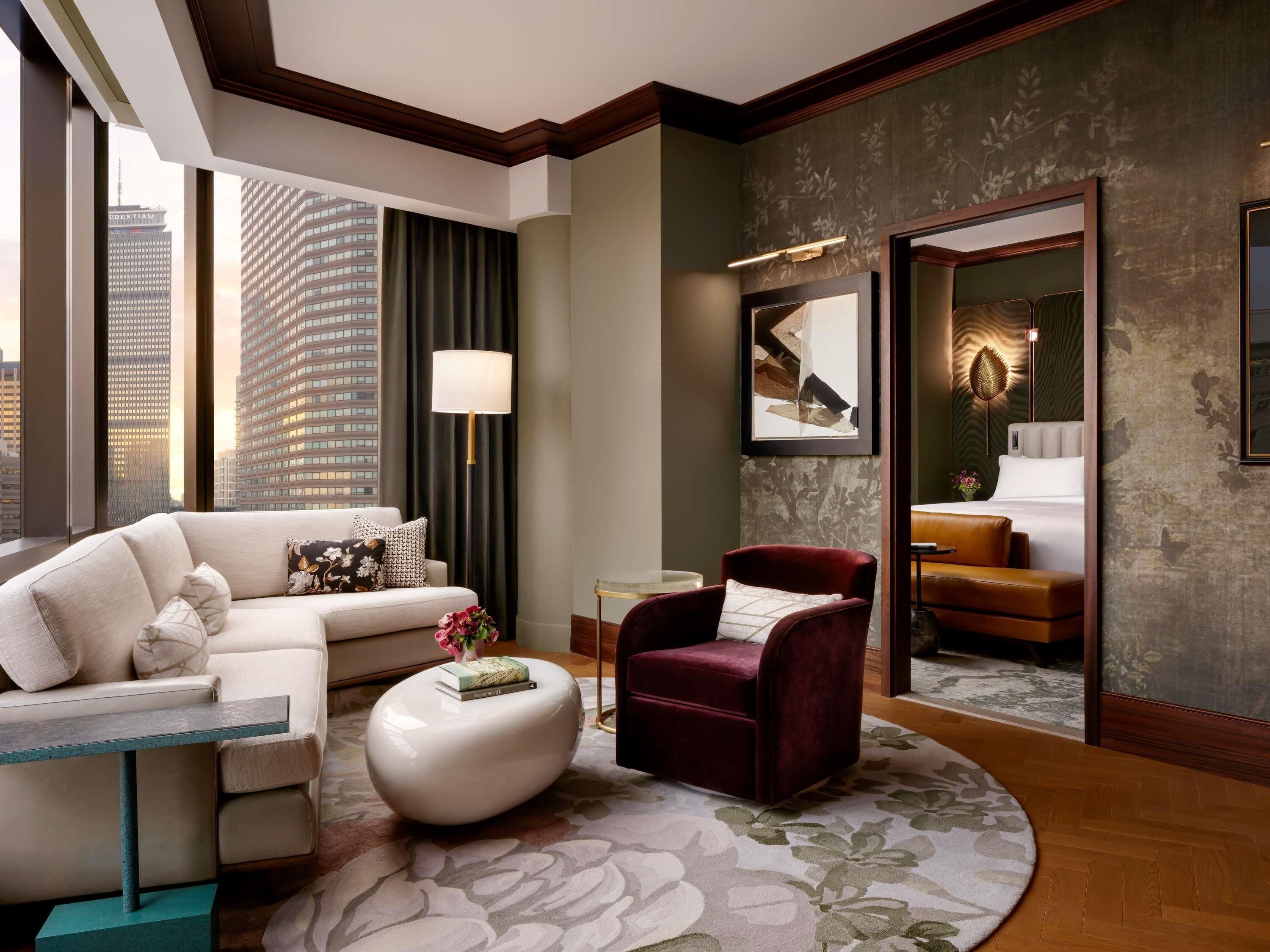 A modern hotel room with a large window, a sofa, a red armchair, a round coffee table, and a bed with a view of the cityscape.