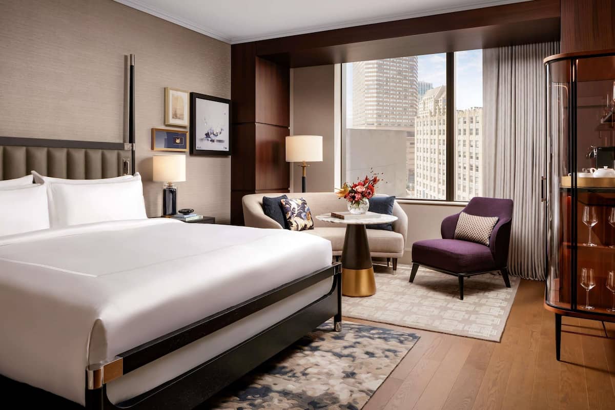 A hotel room with a large bed, a sofa, a chair, a small table with a vase, and a view of the cityscape.