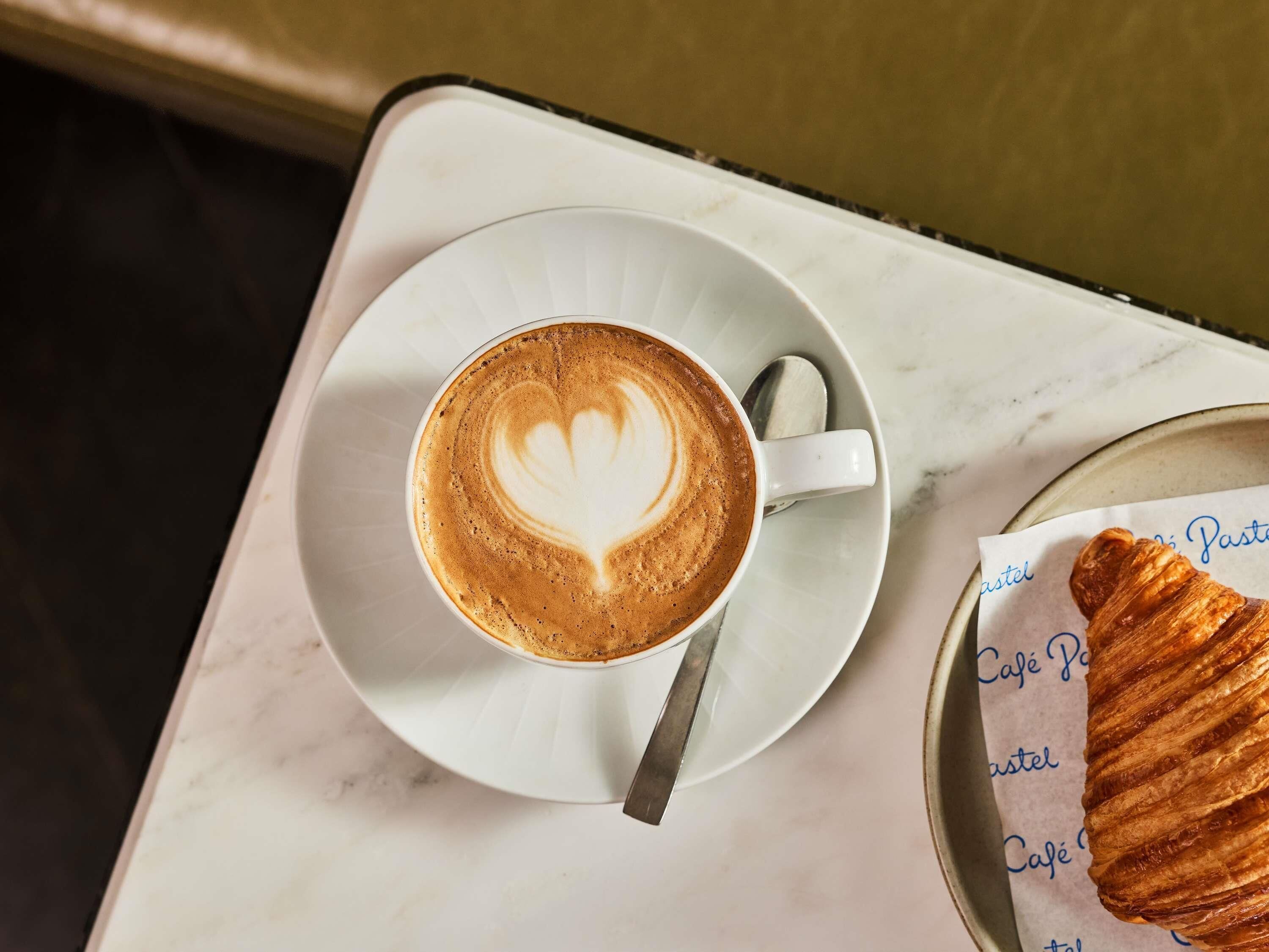 A cup of coffee with latte art on a white saucer, a spoon, and a plate with a croissant on a marble table.