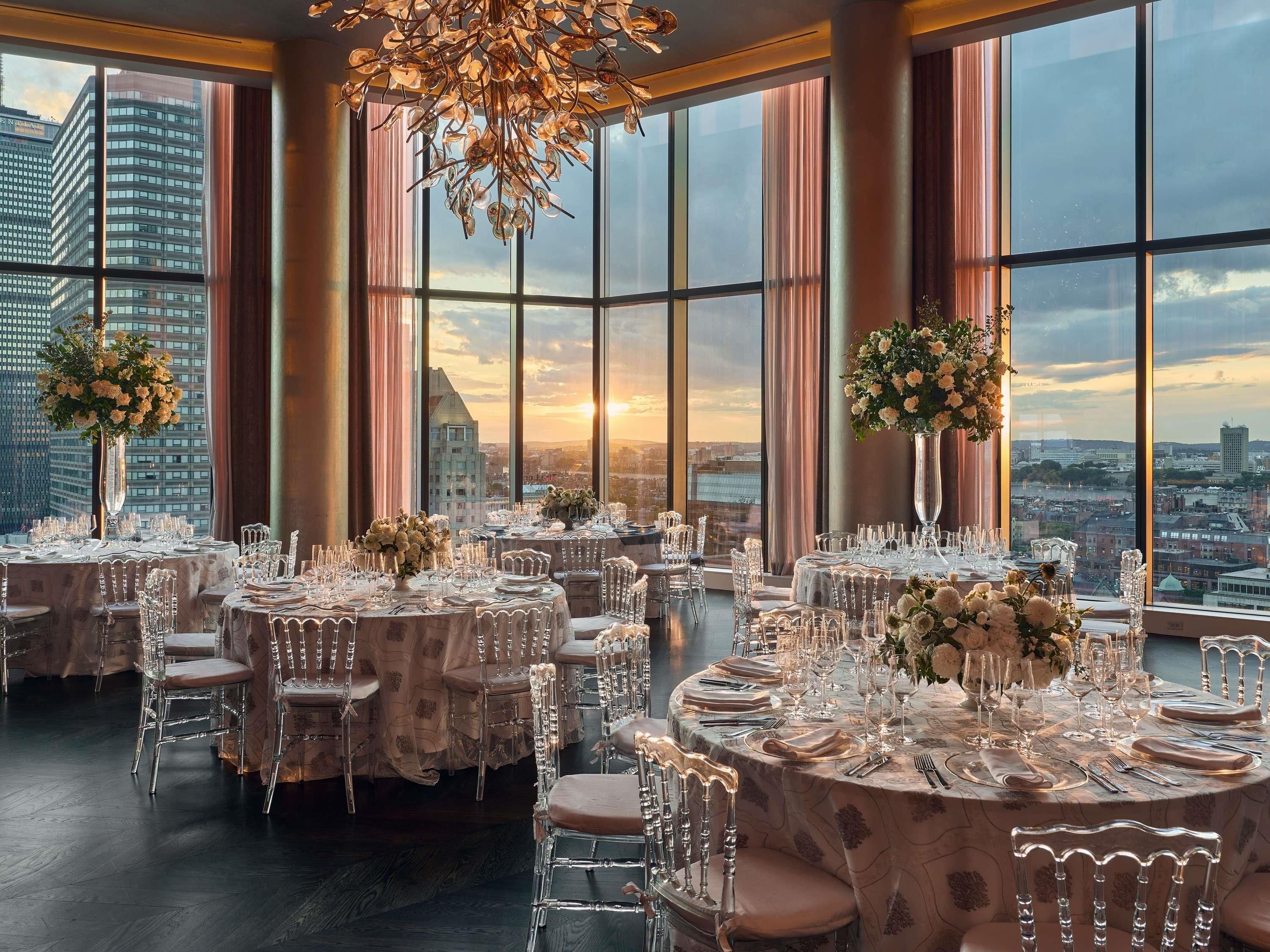 Un comedor en un edificio alto con mesas redondas, sillas transparentes y amplias ventanas con vistas a la ciudad.