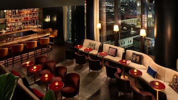 Zona de bar con una barra alta, diversas opciones de asiento como sofás y sillas, y un amplio ventanal con vista a la ciudad.