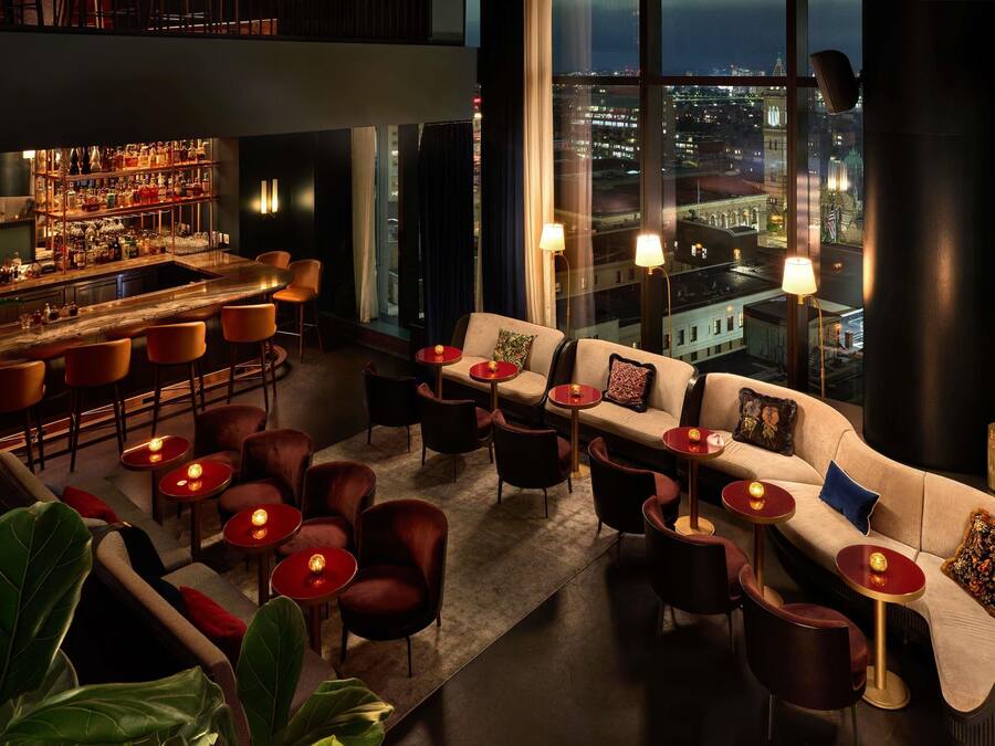 Zona de bar con una barra alta, diversas opciones de asiento como sofás y sillas, y un amplio ventanal con vista a la ciudad.