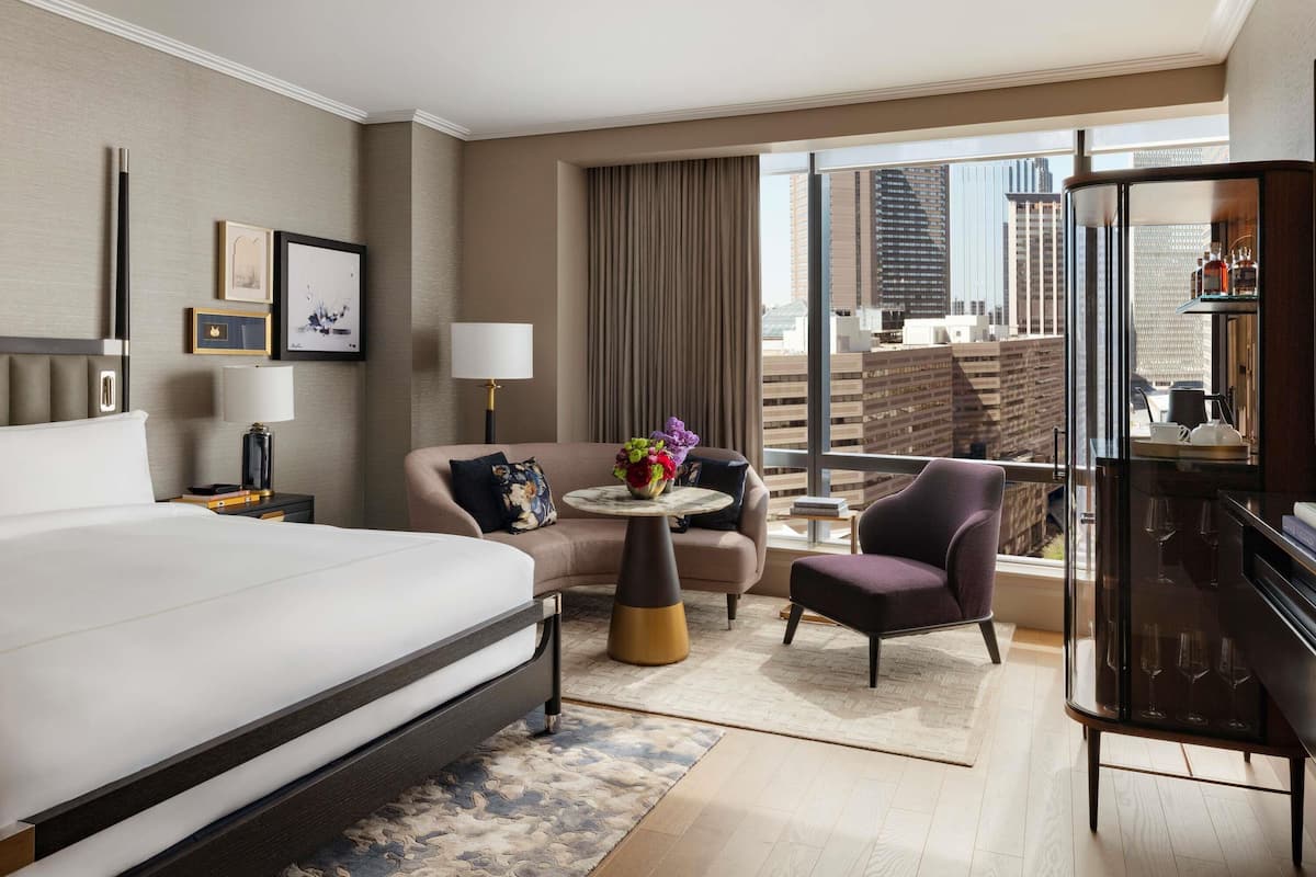 A hotel room with a bed, a desk, a chair, a small table with a vase of flowers, and a view of the city through large windows.