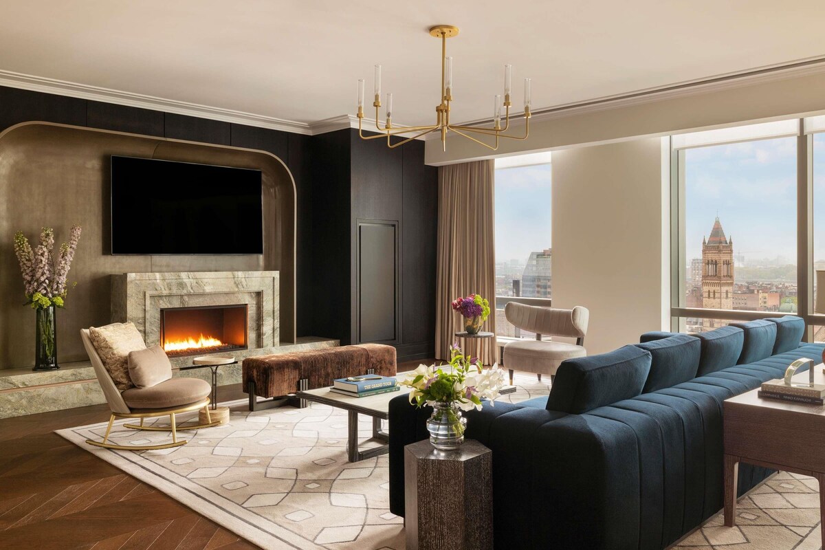 A modern living room with a fireplace, comfortable seating, a large window with a city view, and a chandelier.