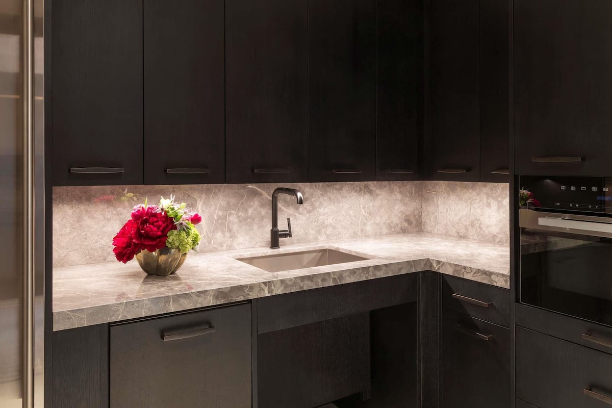 A modern kitchen with dark cabinetry, a marble countertop, a stainless steel sink, and a built-in oven.