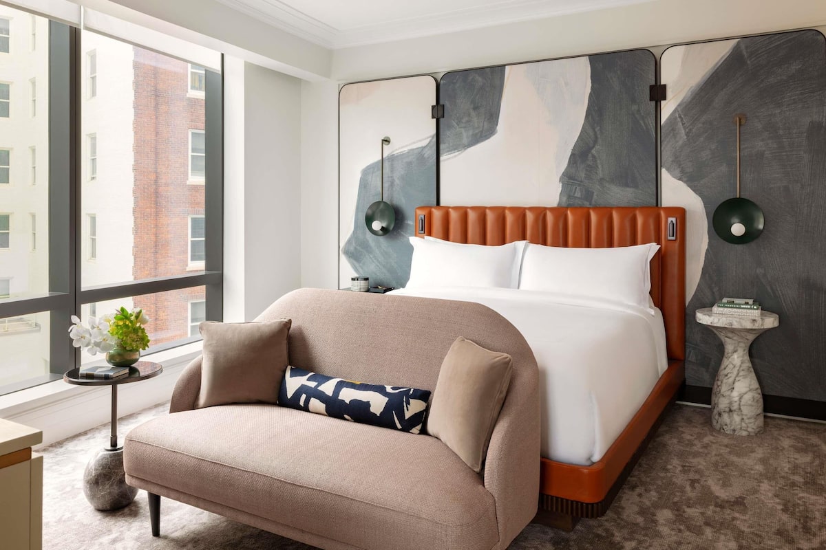 Una habitación de hotel moderna con una cama grande, un sofá, una mesita auxiliar y un ventanal con vistas a la ciudad.