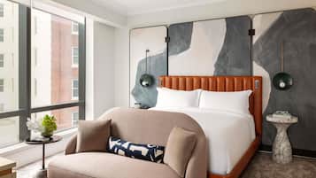 A modern hotel room with a large bed, a sofa, a small side table, and a large window offering a view of the city.