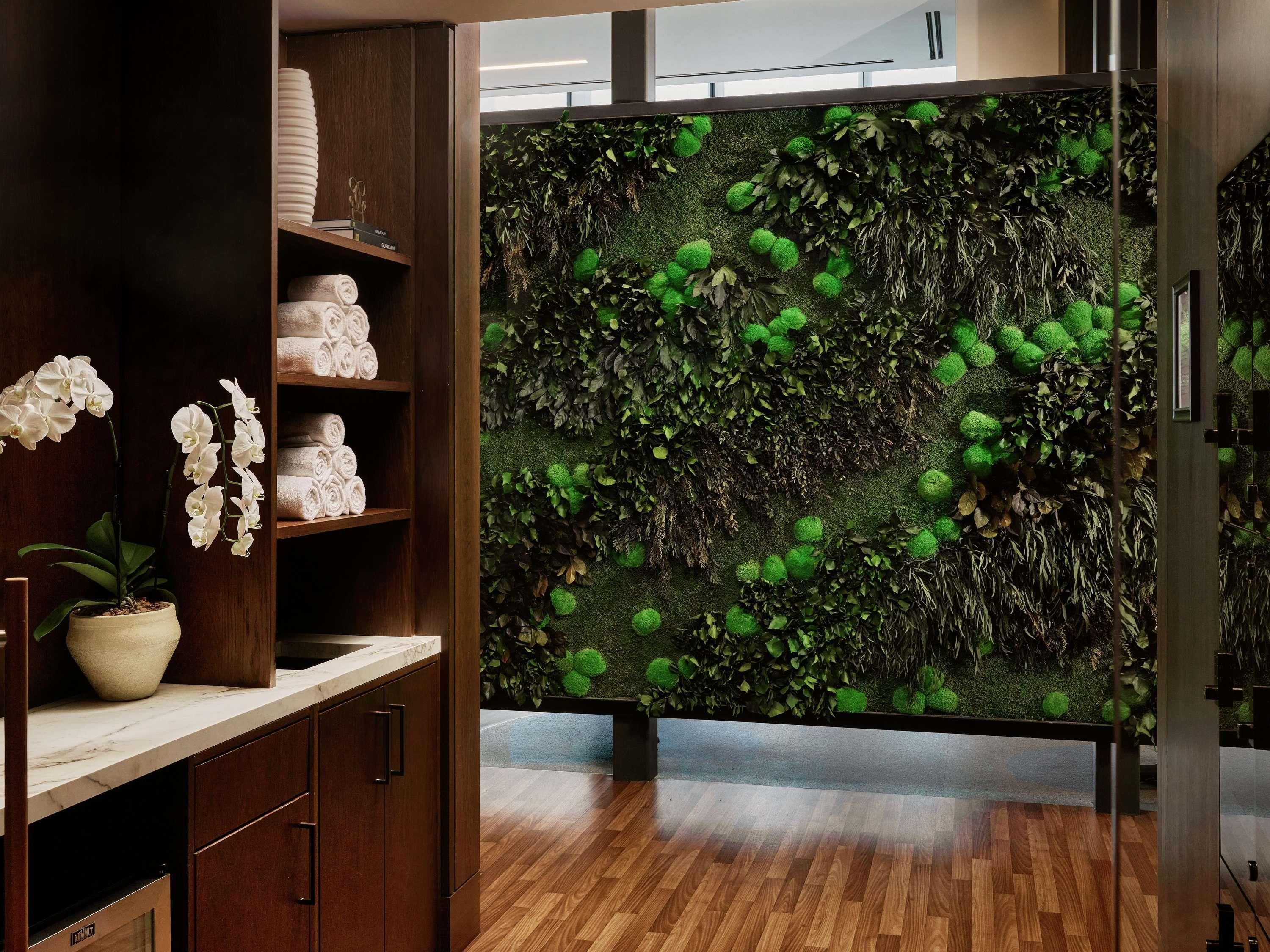 Un interior moderno con un estante de madera que sostiene toallas dobladas, una orquídea en maceta y un diseño de musgo y hojas en la pared.