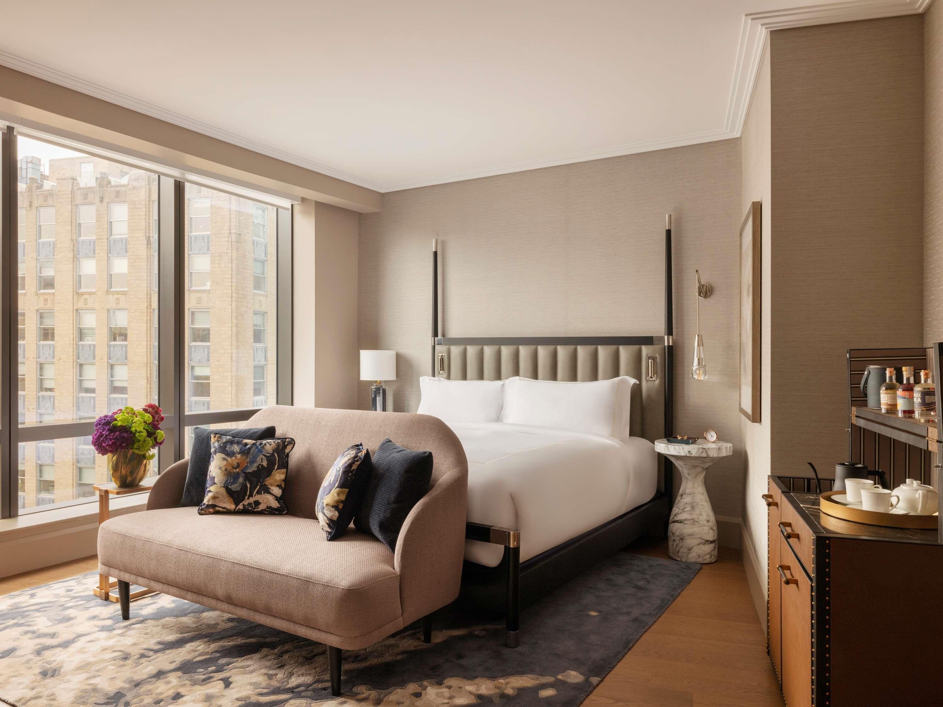 A hotel room with a bed, a sofa, a side table, a lamp, a vase with flowers, a window with a view of buildings, and a minibar.