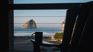 A chair by a window overlooking a beach with a rock formation, a car, and a building. - Inn at Cape Kiwanda (Pacific City)