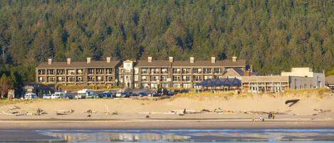 Un hôtel situé sur la côte, composé de plusieurs bâtiments, avec une plage au premier plan et une colline boisée en arrière-plan.