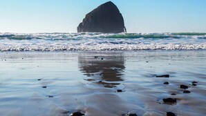 A beach with a large rock formation in the water, waves gently crashing onto the shore, and a clear sky. - Inn at Cape Kiwanda (Pacific City)