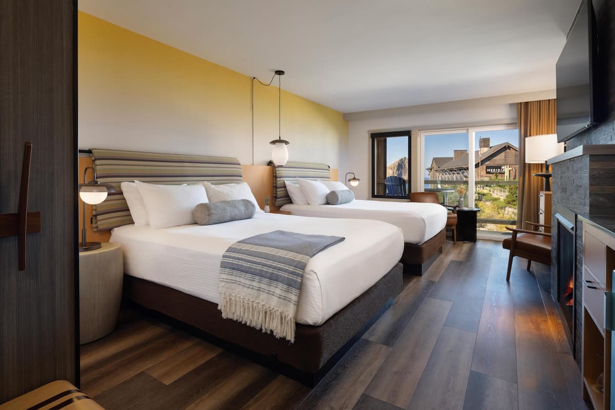A modern hotel room with two beds, a TV, a fireplace, and a balcony with a view of the mountains.