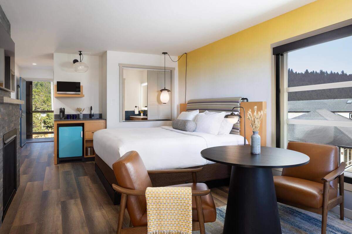 A modern hotel room with a large bed, a dining table, and a view of the outdoors.