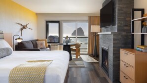 A hotel room with a large bed, a sofa, a dining table, a fireplace, and a TV. The room has a view of the ocean and a mountain. - Inn at Cape Kiwanda (Pacific City)