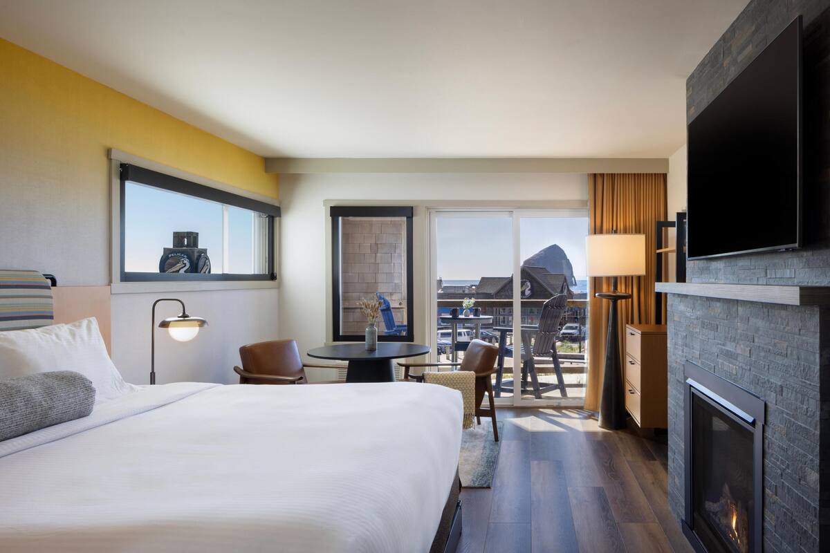 A hotel room with a large bed, a desk with a chair, a fireplace, a TV mounted on the wall, and a balcony with a view.