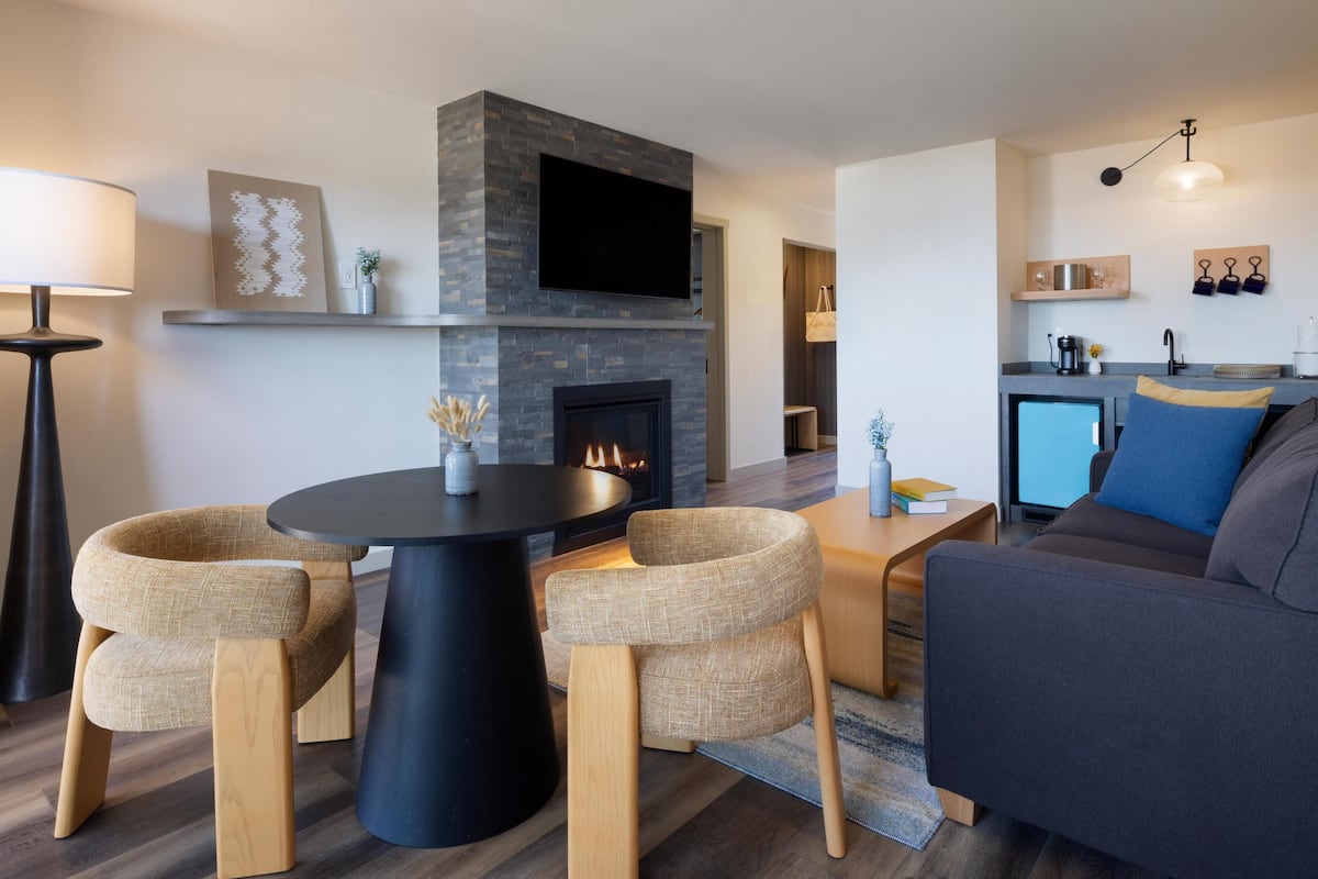 A modern living room with a fireplace, a flat-screen TV mounted above it, a round coffee table, two armchairs, a sofa, and a small kitchen area with a sink and shelves.