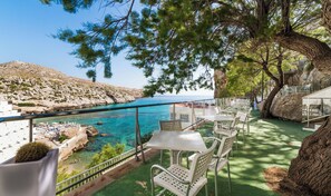 A terrace with outdoor seating overlooking a coastal area with clear blue water and rocky terrain.