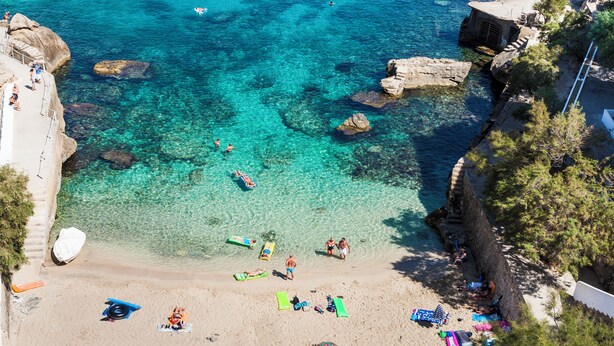 Ein Strand mit klarem türkisfarbenem Wasser, Felsformationen und Menschen, die sich sonnen und schwimmen.