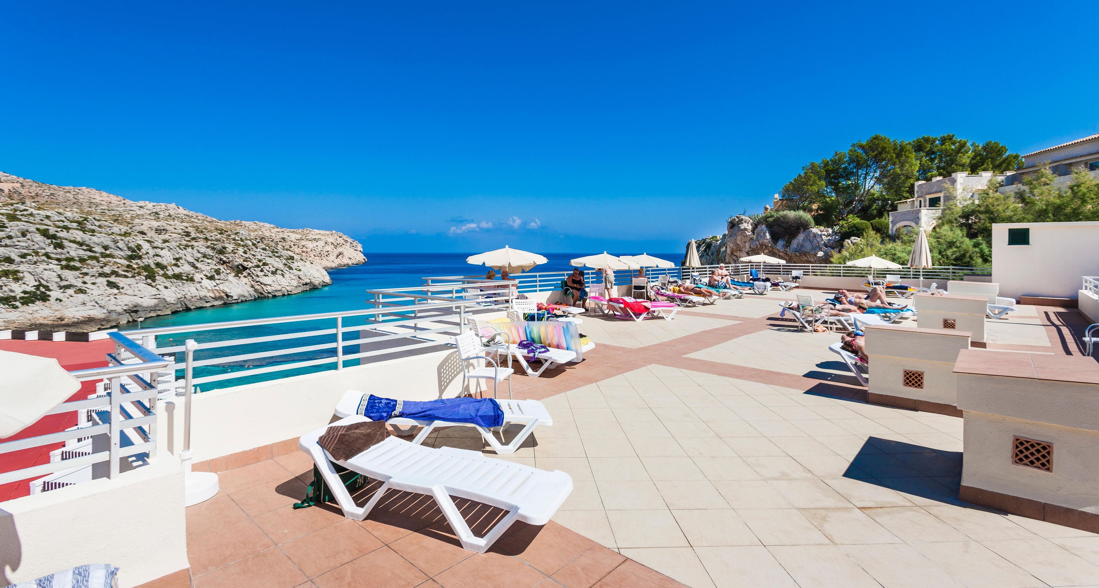 A sunlit outdoor seating area with lounge chairs and umbrellas, overlooking a clear blue sea and rocky coastline.