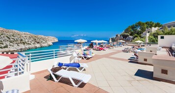 Un espacio al aire libre con sol, sillones de descanso y sombrillas, con vistas al mar azul y a la costa rocosa.
