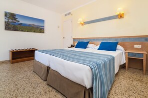 Hotellihuone, jossa on sänky, yöpöytä, sängyn vieressä oleva pöytä, seinään kiinnitetty lamppu ja maisemakuva.