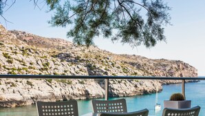 Eine Terrasse mit drei Metallstühlen und einem Tisch, mit Blick auf eine felsige Küstenlinie und klares blaues Meer.
