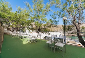 An outdoor seating area with tables and chairs, surrounded by trees and overlooking a body of water. - Globales Simar (Pollença)