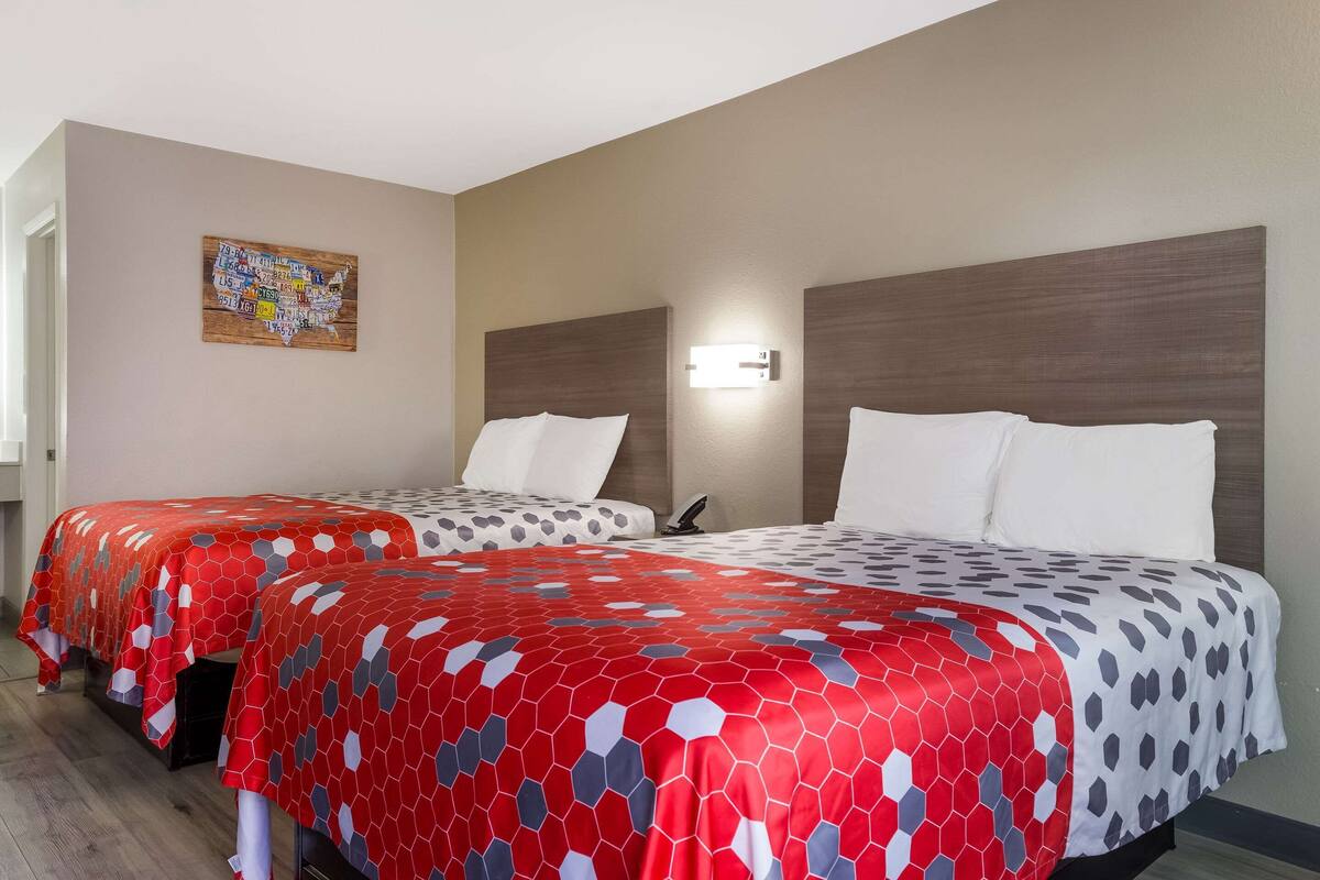 A hotel room with two beds, each with a red and gray patterned bedspread, a wooden headboard, a wall-mounted light, and a map artwork on the wall.