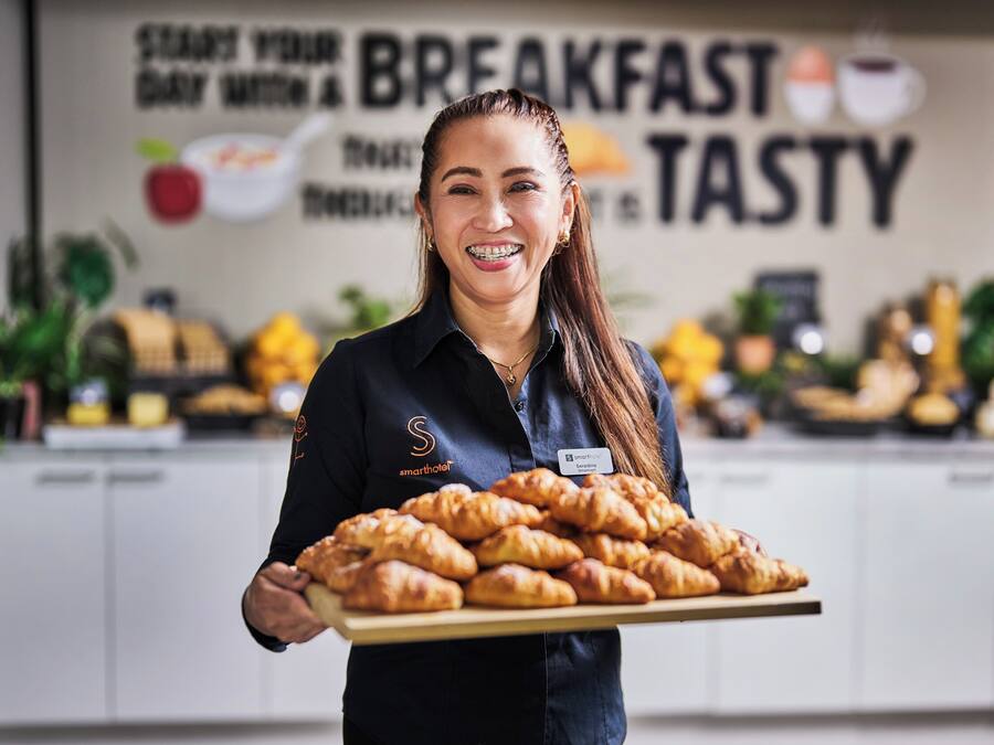Una persona sostiene una bandeja de cruasanes en una cocina con un cartel que dice 'EMPIEZA EL DÍA CON UN DESAYUNO DELICIOSO'.