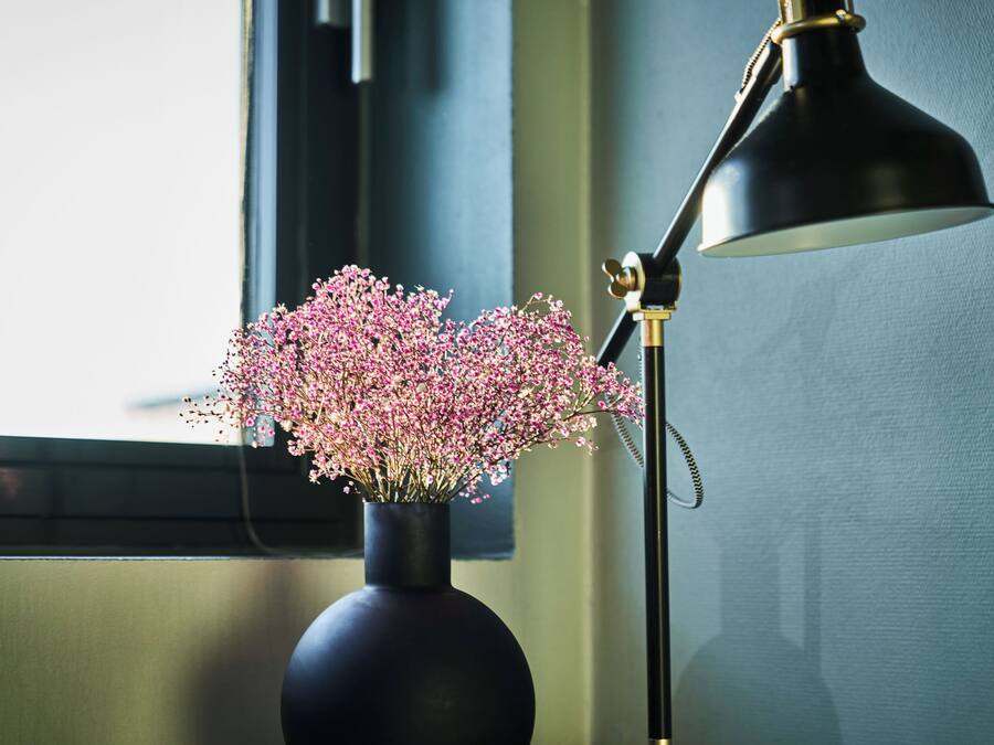 Eine Vase mit rosa Blumen auf einem Fensterbrett neben einer schwarzen Schreibtischlampe.