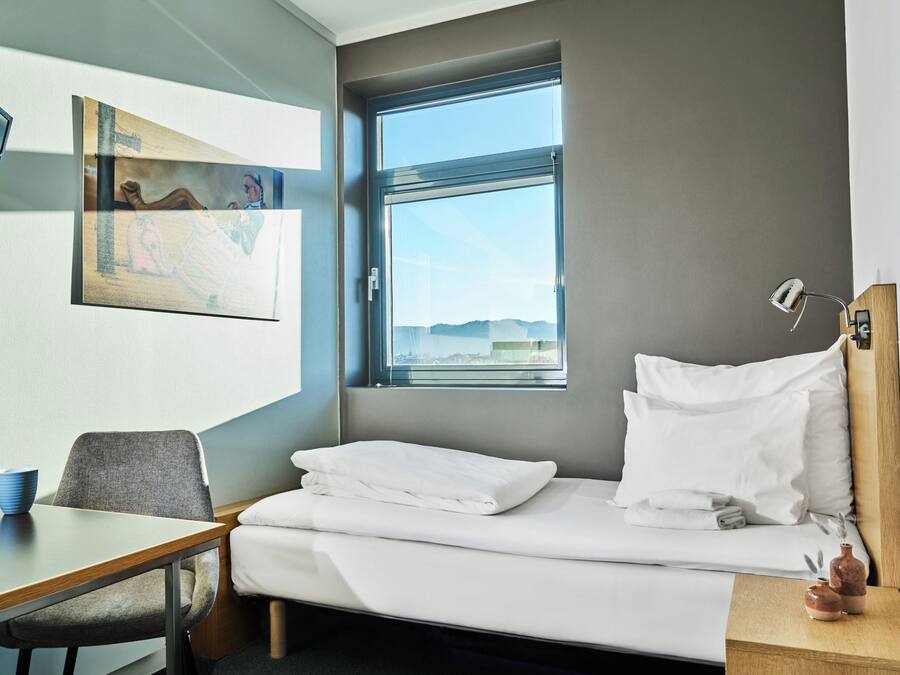 A hotel room with a bed, a desk, a chair, a window with a view, and a painting on the wall.