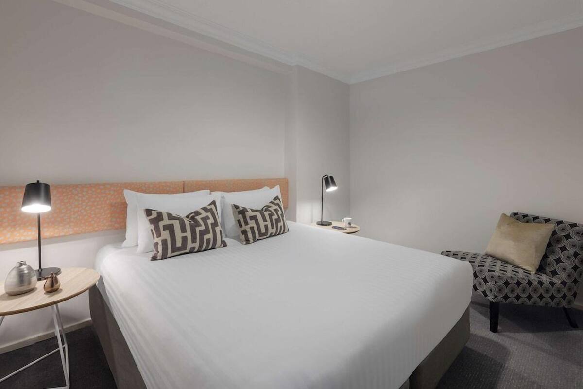 A hotel room with a large bed, two bedside tables, a patterned armchair, and a lamp.
