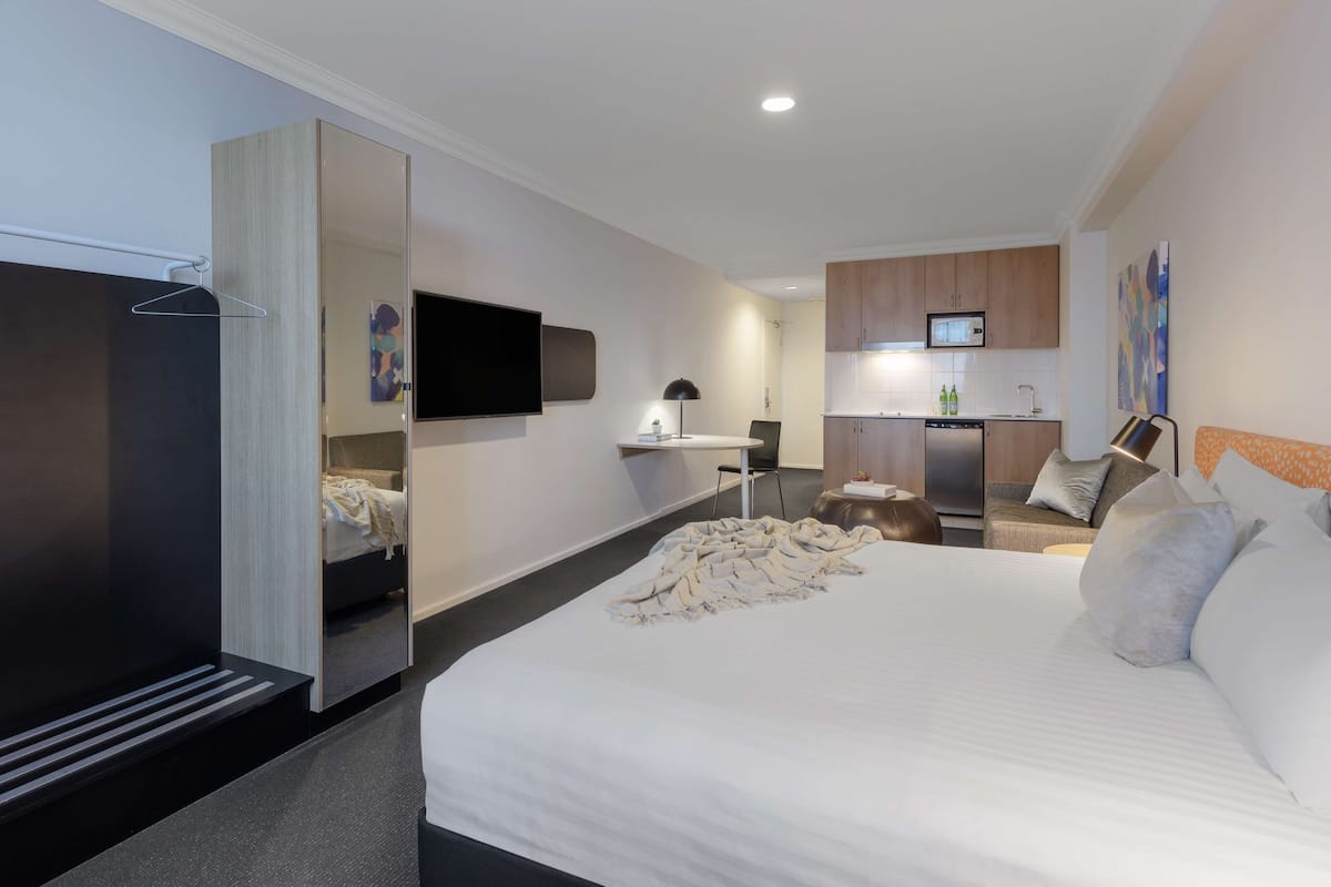 A modern hotel room with a large bed, a desk, a flat-screen TV, and a kitchenette.