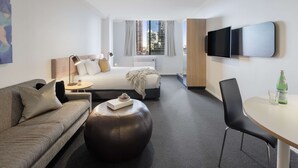 A modern hotel room with a bed, a sofa, a round coffee table, a TV mounted on the wall, and a window with curtains. - Oaks Perth Hotel (Perth)