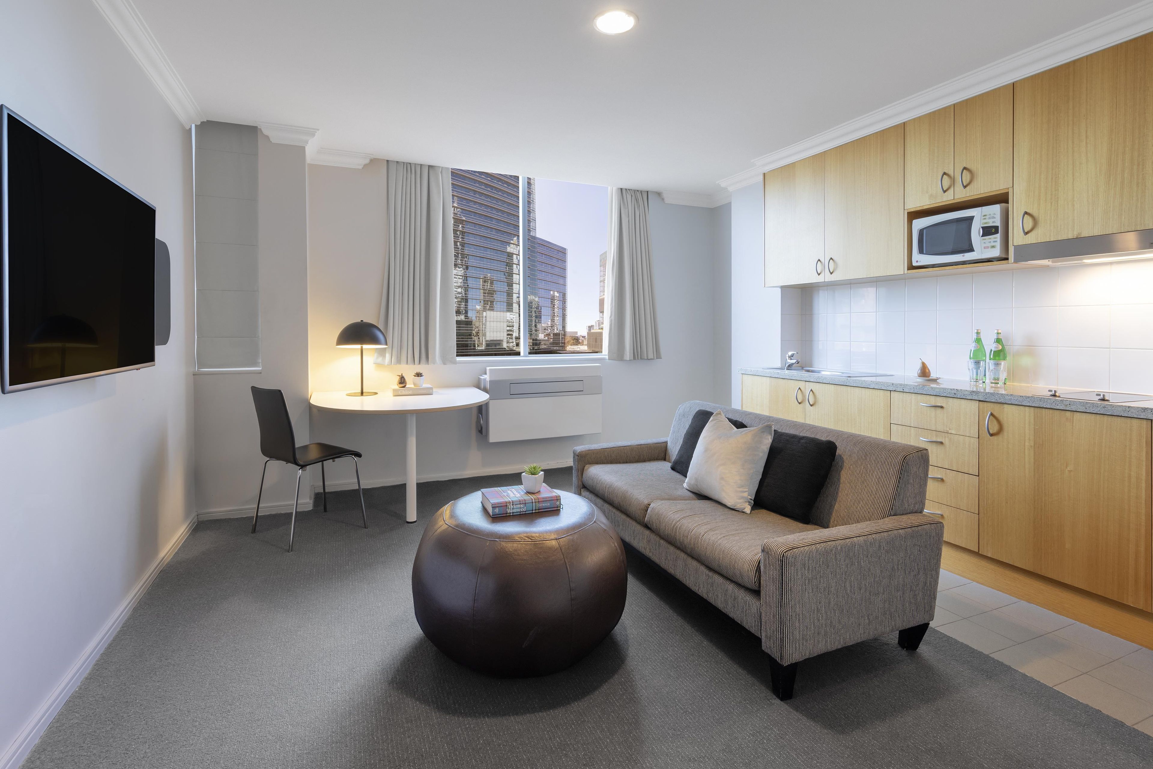A modern living room with a sofa, a round ottoman, a desk, and a flat-screen TV.