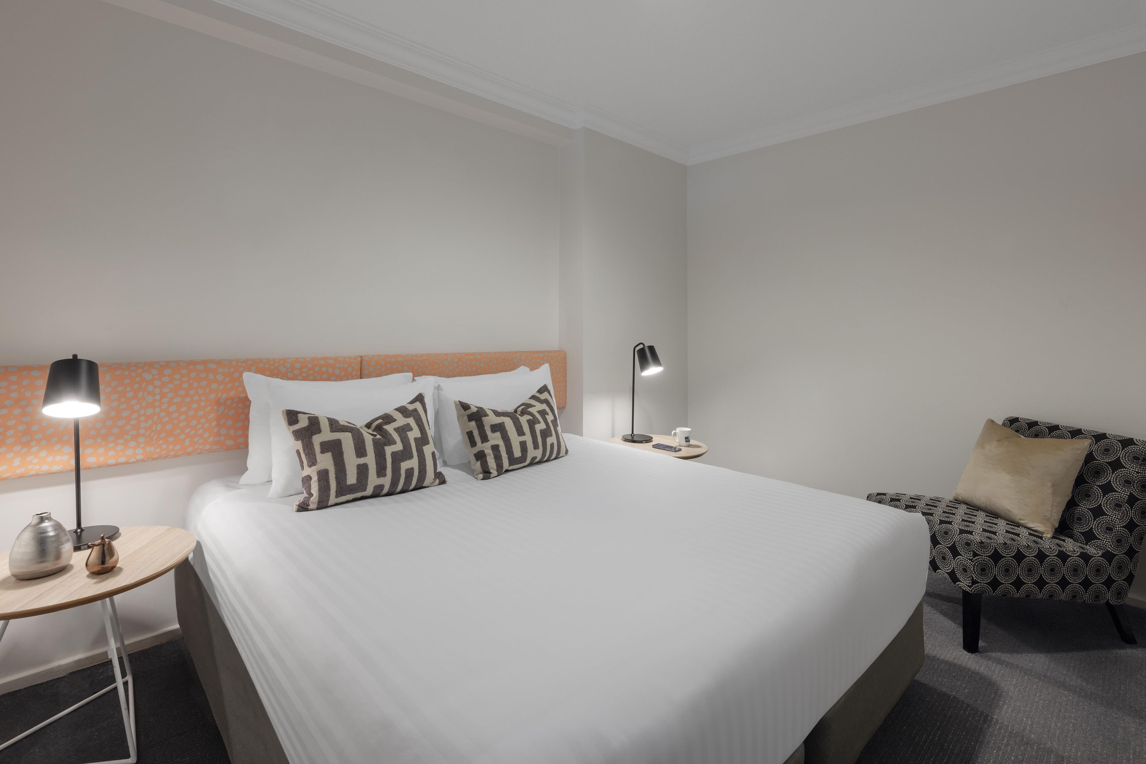 A hotel room with a large bed, two bedside tables, a nightstand, a lamp, a chair, and a coffee table.