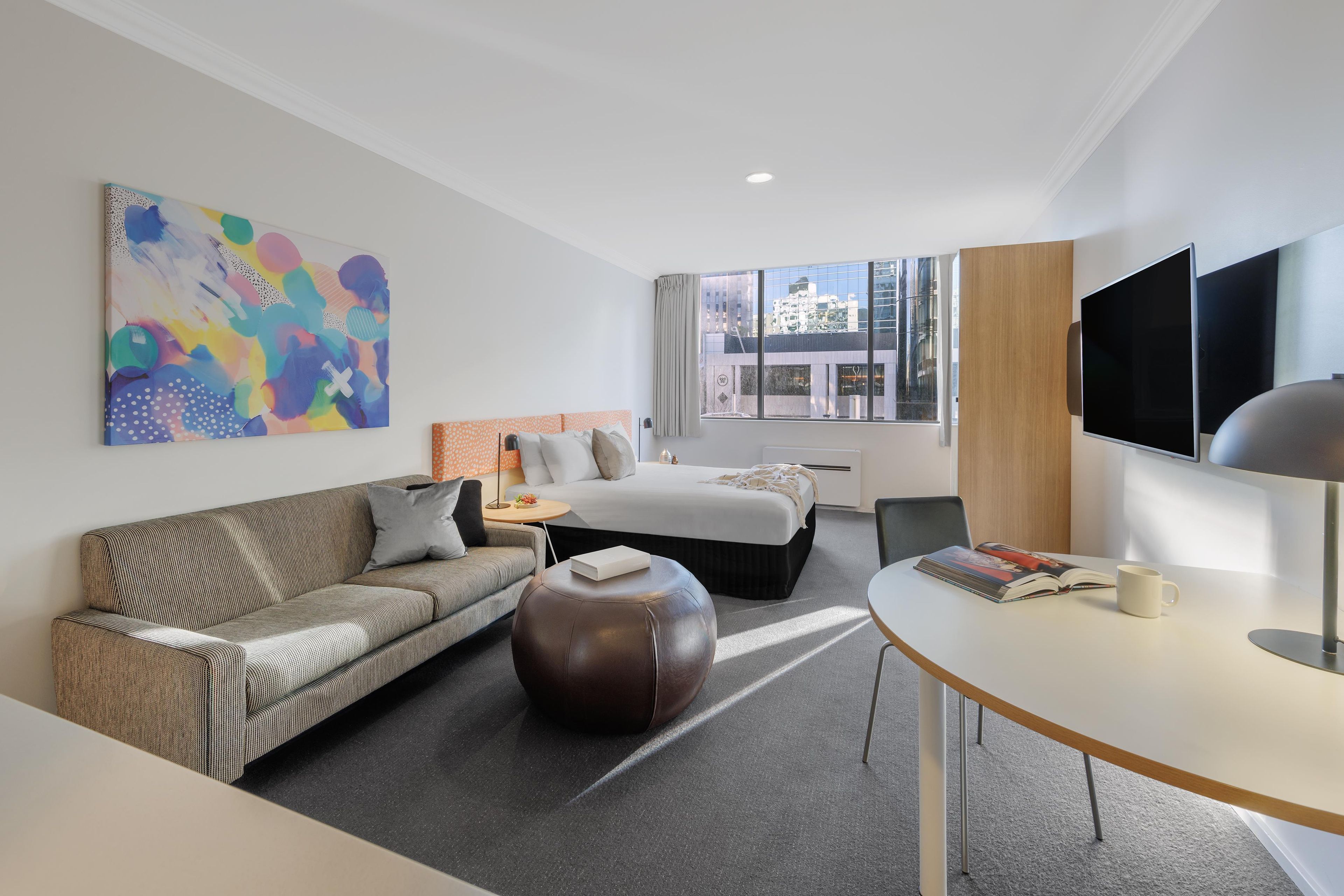 A modern hotel room with a sofa, a bed, a dining table, and a TV.