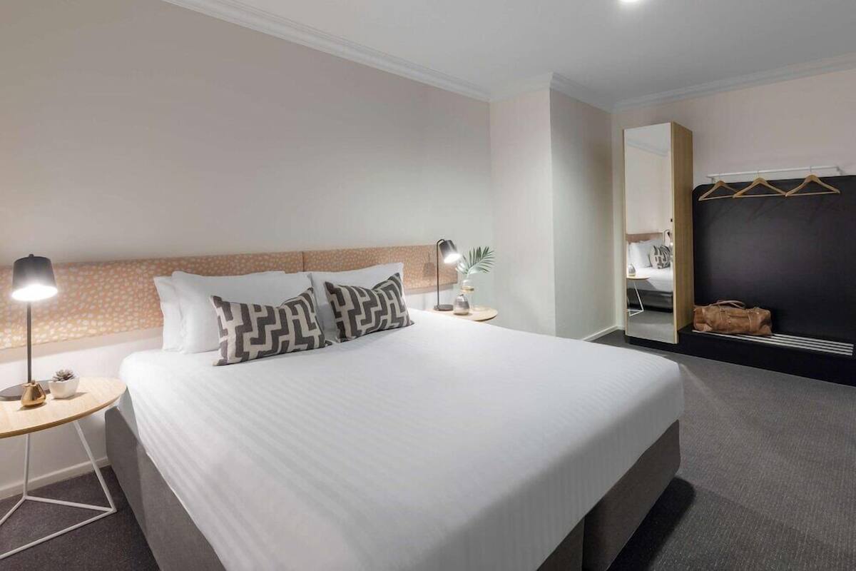 A neatly made bed with patterned pillows, a bedside lamp, a small table with a plant, and a mirror with a hanging bag.