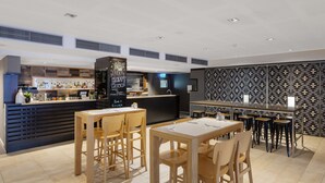 A modern bar area with wooden tables and chairs, a bar counter with a menu, and a patterned wall in the background. - Oaks Perth Hotel (Perth)