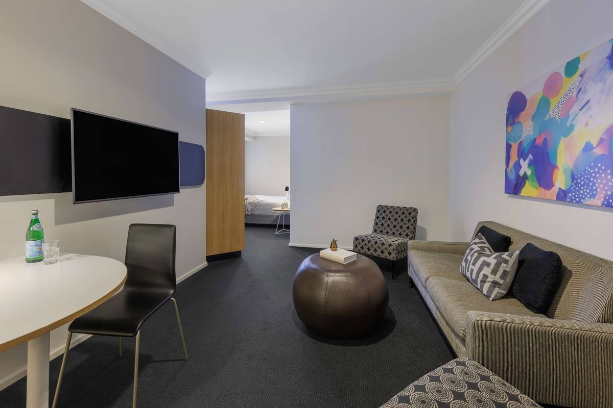 A modern hotel room with a flat-screen TV, a round ottoman, a sofa, and a colorful abstract painting.