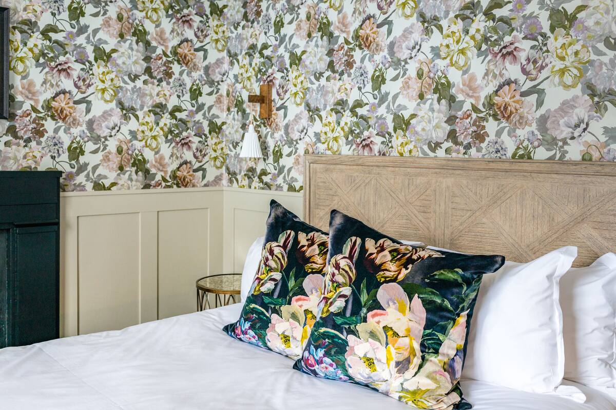 A bedroom with a floral wallpaper, a wooden headboard, and two decorative pillows.